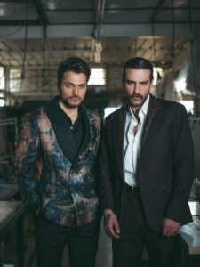 Two men in artistic suits pose confidently in an industrial indoor setting.